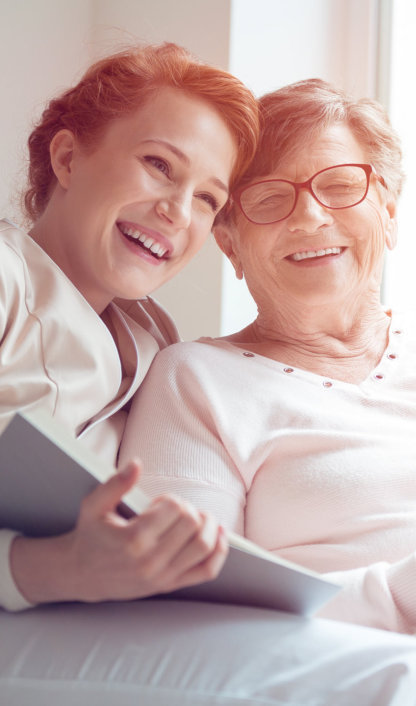 Young caregiver and senior women laughing together while sitting on sofa